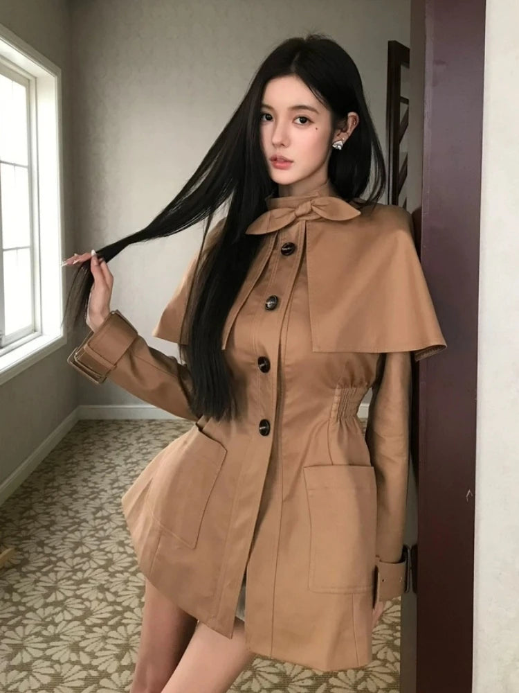 Woman in a brown coat standing indoors near a window.