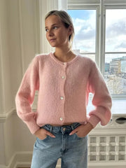 Woman wearing a pink cardigan and blue jeans standing in a room with large windows.