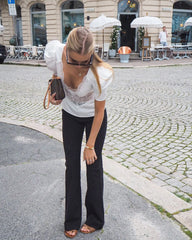 Woman in white blouse and black pants walking on a street with buildings and tables in the background.