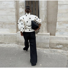 Person wearing a white polka dot shirt and black pants walking past a stone building.