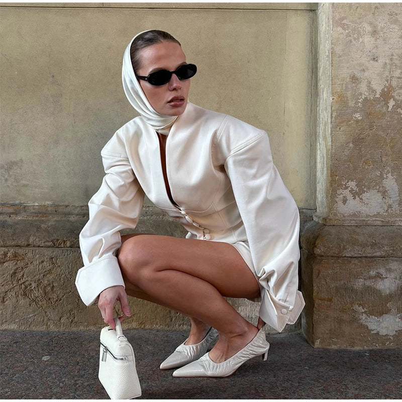 Woman in a white outfit with a headscarf and sunglasses, posing against a textured wall.
