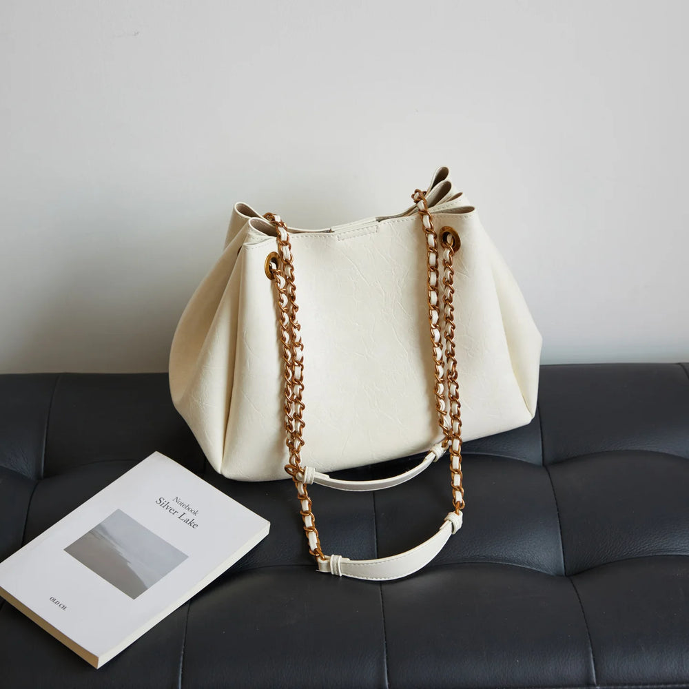 White handbag with gold chain handle on a black leather couch next to a book.