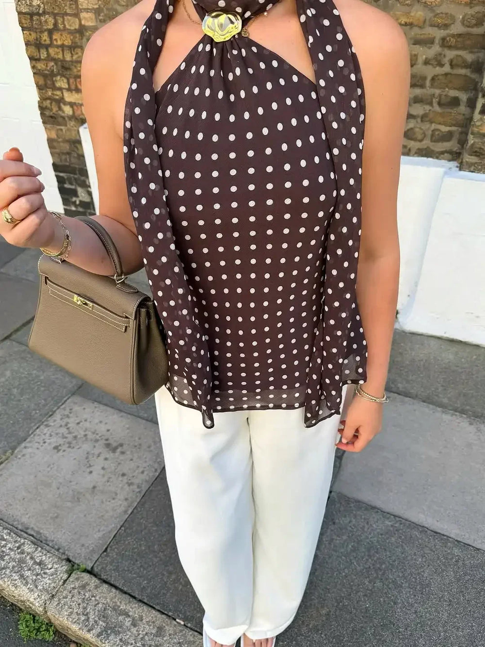 Woman wearing a brown polka dot scarf, white pants, and holding a beige handbag on a street.