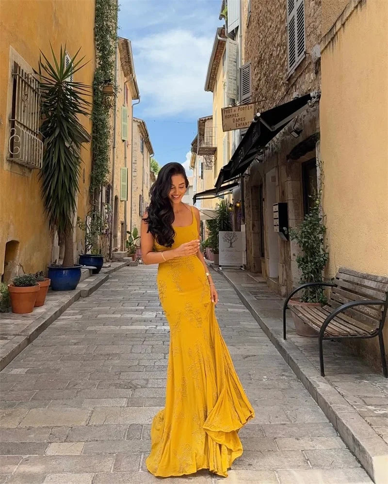 Woman in a yellow dress standing on a narrow street with buildings and plants.
