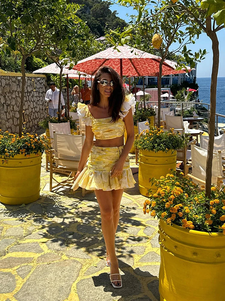 Woman in a yellow dress standing in a scenic outdoor setting with flowers and tables.