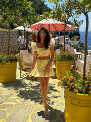 Woman in a yellow dress standing in a scenic outdoor setting with flowers and tables.