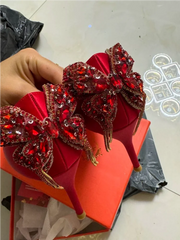Red high-heeled shoes with decorative elements held by a hand on a light-colored floor.
