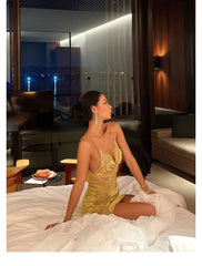 Woman in a gold dress sitting on a bed in a luxurious room with city lights visible through the window.