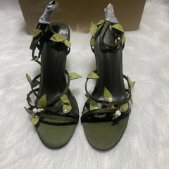 Pair of black high-heeled sandals with green leaf-like straps and rhinestone embellishments on a white textured surface.