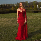 Model wearing a red crystal-embellished one-shoulder trumpet gown in a garden setting.