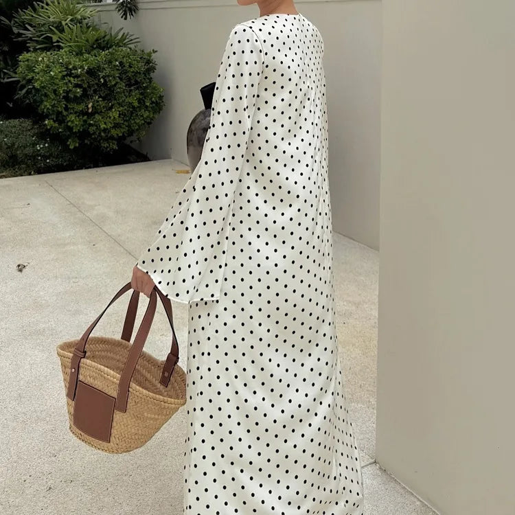 Person wearing a long white dress with black polka dots, holding a woven bag.