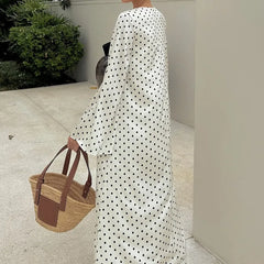 Person wearing a long white dress with black polka dots, holding a woven bag.