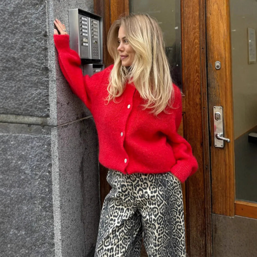 Woman in a red sweater and leopard print pants leaning against a building wall.