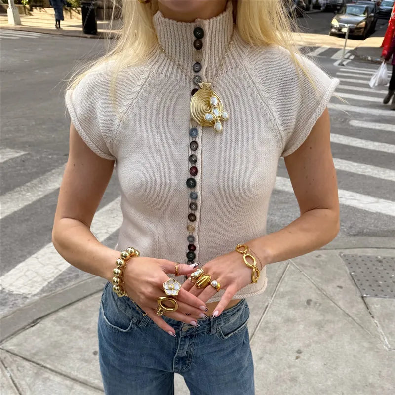 Person wearing a sleeveless knit top with decorative buttons and holding jewelry on a city street.