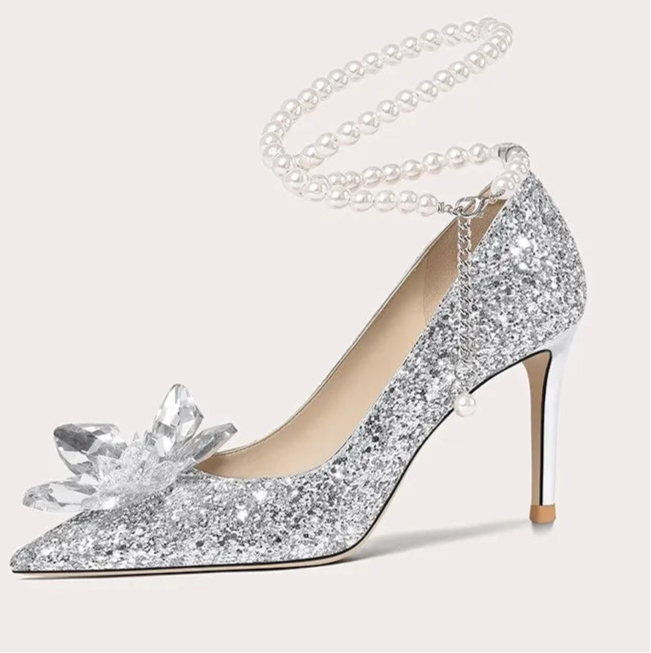 Silver glitter high-heeled shoe with a decorative bow and pearl chain on a white background