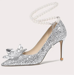Silver glitter high-heeled shoe with a decorative bow and pearl chain on a white background