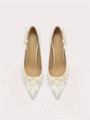 Pair of white pointed-toe shoes with floral embellishments on a light gray background