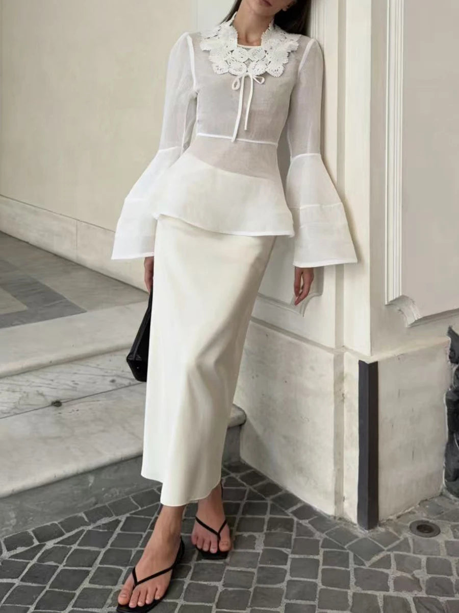 Woman in a white outfit with a floral collar standing against a neutral wall.