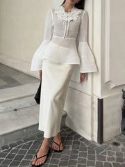 Woman in a white outfit with a floral collar standing against a neutral wall.