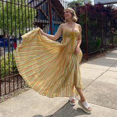 Woman in a yellow striped dress standing on a sidewalk with a fence and trees in the background.