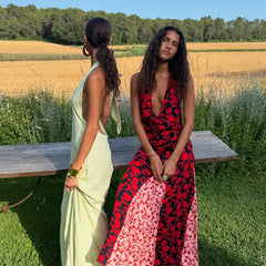 Two women sitting on a wooden bench in a field with a cornfield and trees in the background.