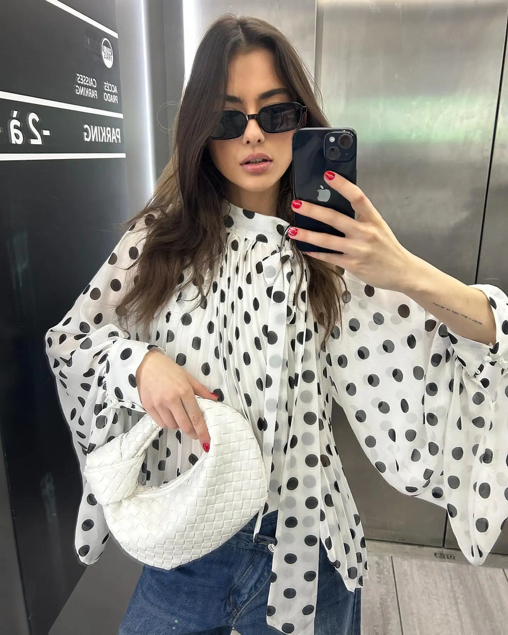 Woman taking a mirror selfie wearing a polka dot blouse and sunglasses, holding a phone.