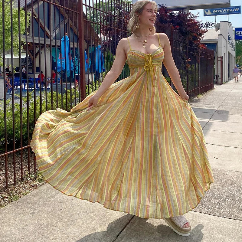 Woman in a yellow striped dress standing on a sidewalk with a background of a fence and trees.