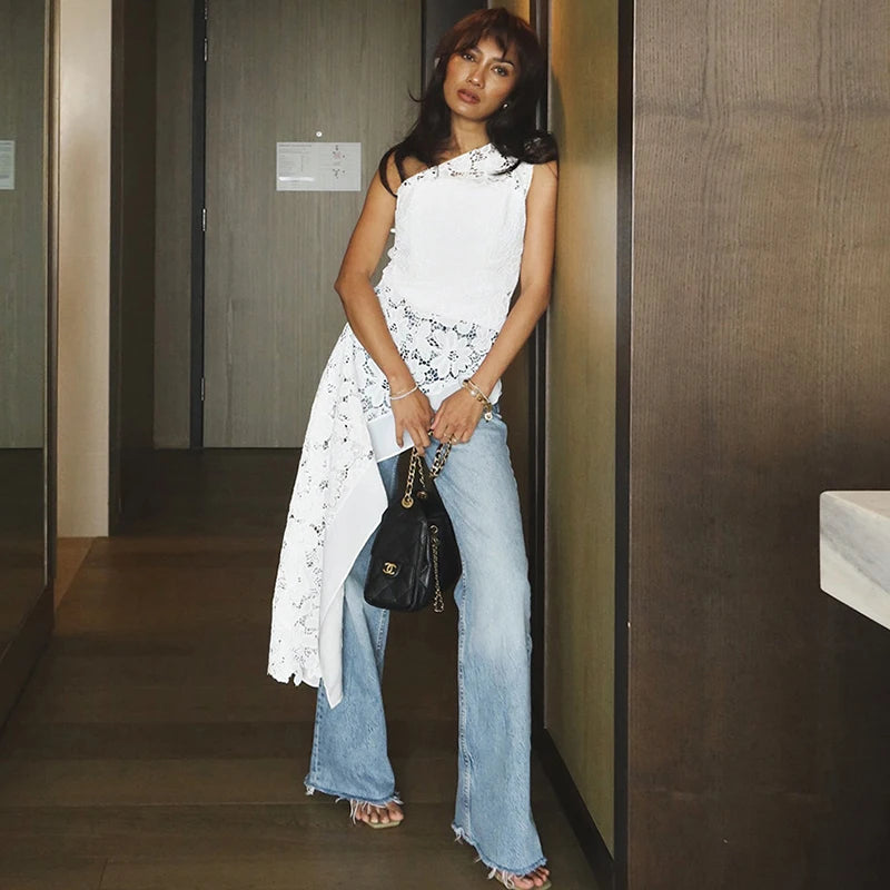 Woman in a white dress and blue jeans holding a black handbag in an indoor setting.