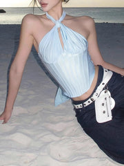 Woman in a light blue halter top and dark pants sitting on sand with ocean in the background