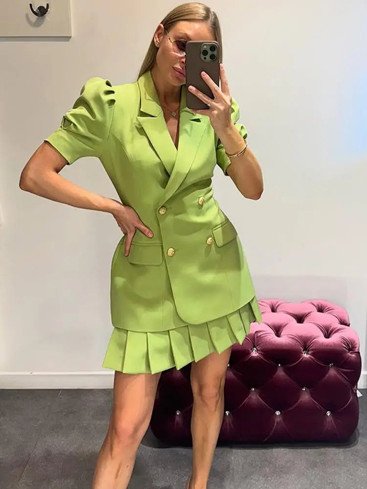 Woman taking a mirror selfie wearing a green suit with a white wall and purple ottoman in the background.