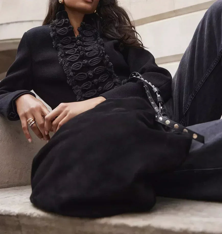 Person wearing a black coat with decorative elements sitting on a bench.