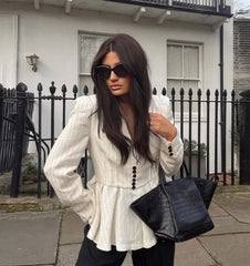 Woman in a white peplum top and black pants holding a black handbag, standing in front of a house.
