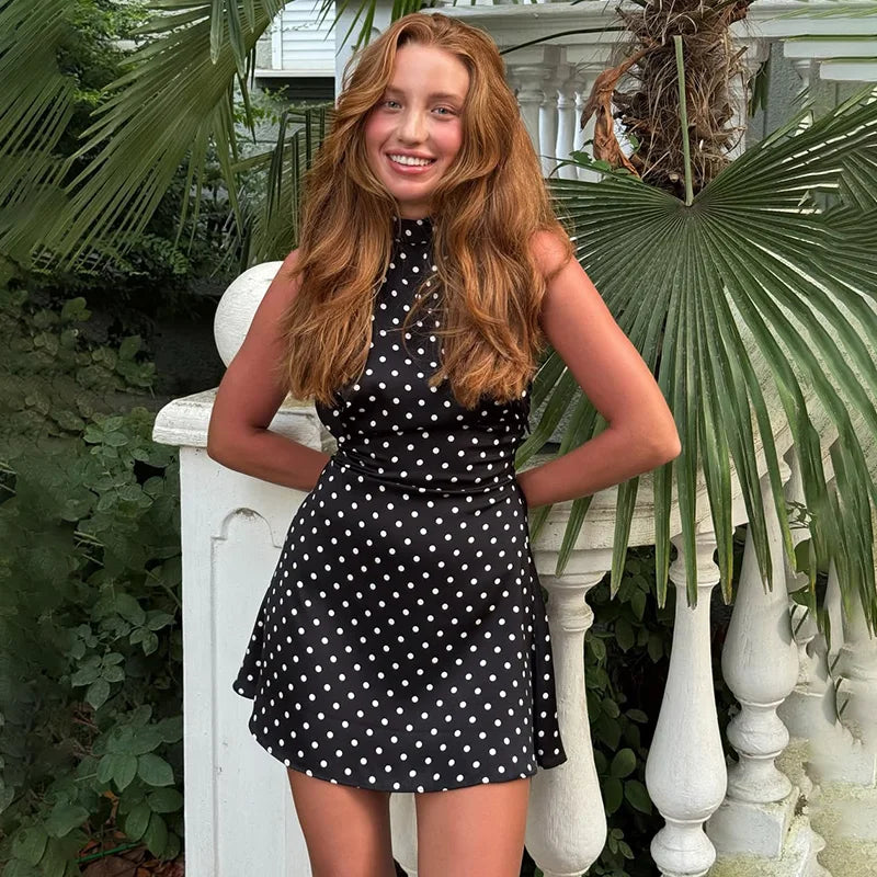 Woman in a black polka dot dress standing outdoors with greenery.