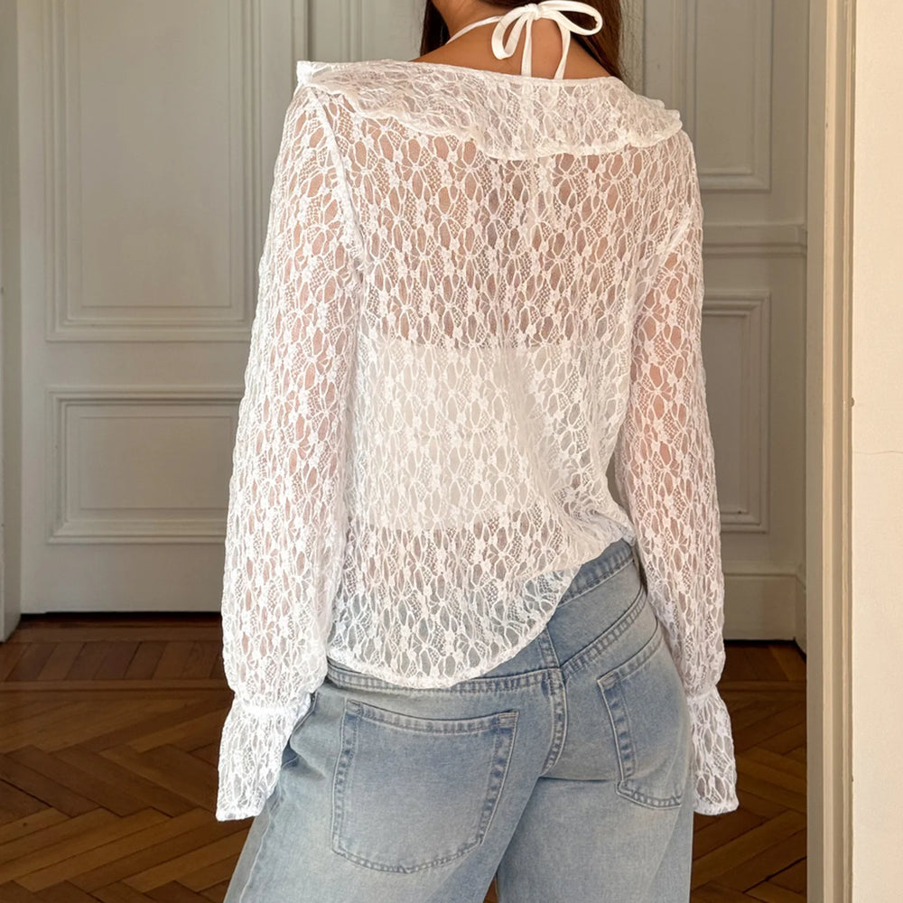 Person wearing a white lace top and light blue jeans in a room with wooden flooring and gray walls.