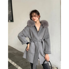 Woman wearing a gray coat with fur hood standing against a plain wall.