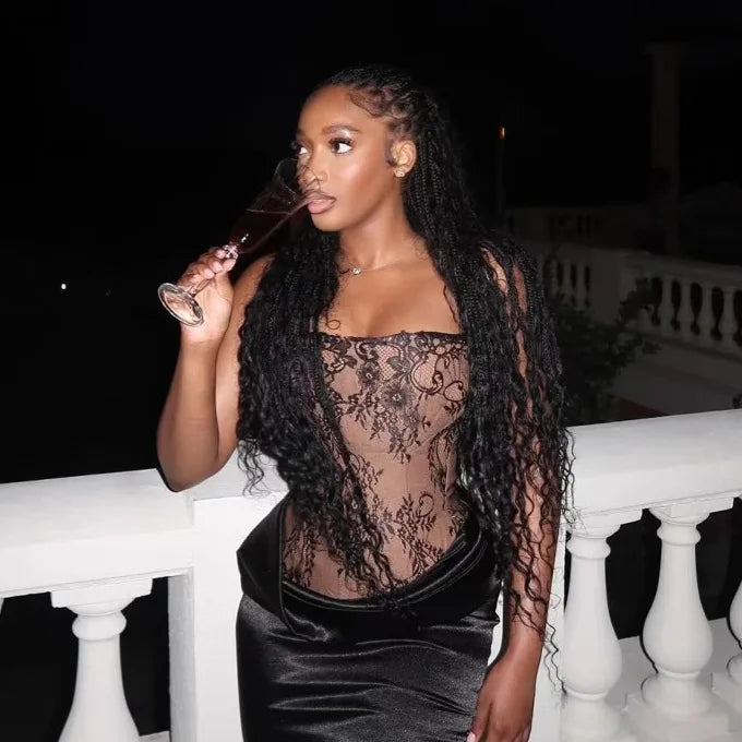 Woman in a sheer black lace top and black pants holding a glass on a balcony at night.