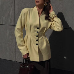 Woman wearing a light yellow blazer with black buttons against a gray wall.