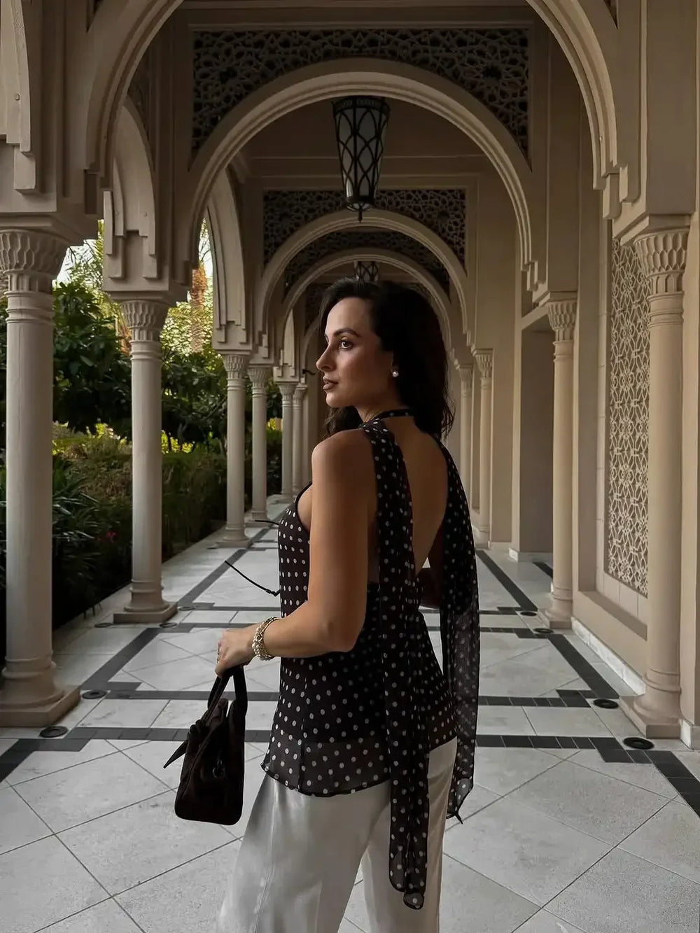 Woman in a black polka dot top and white pants standing in an ornate colonnade.