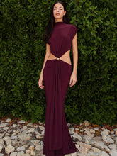 Woman wearing a burgundy evening gown with cut-out design against a green hedge background.
