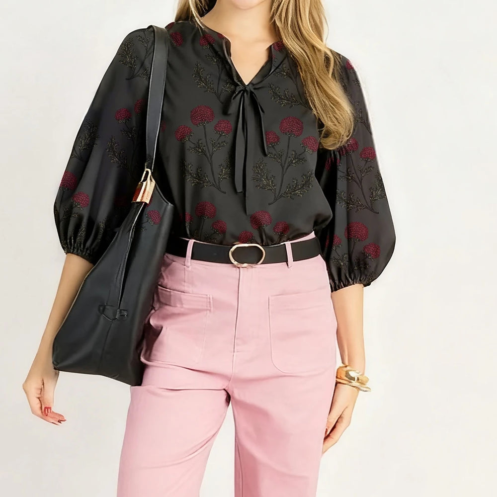 Woman wearing a black floral blouse and pink pants with a black bag on a white background