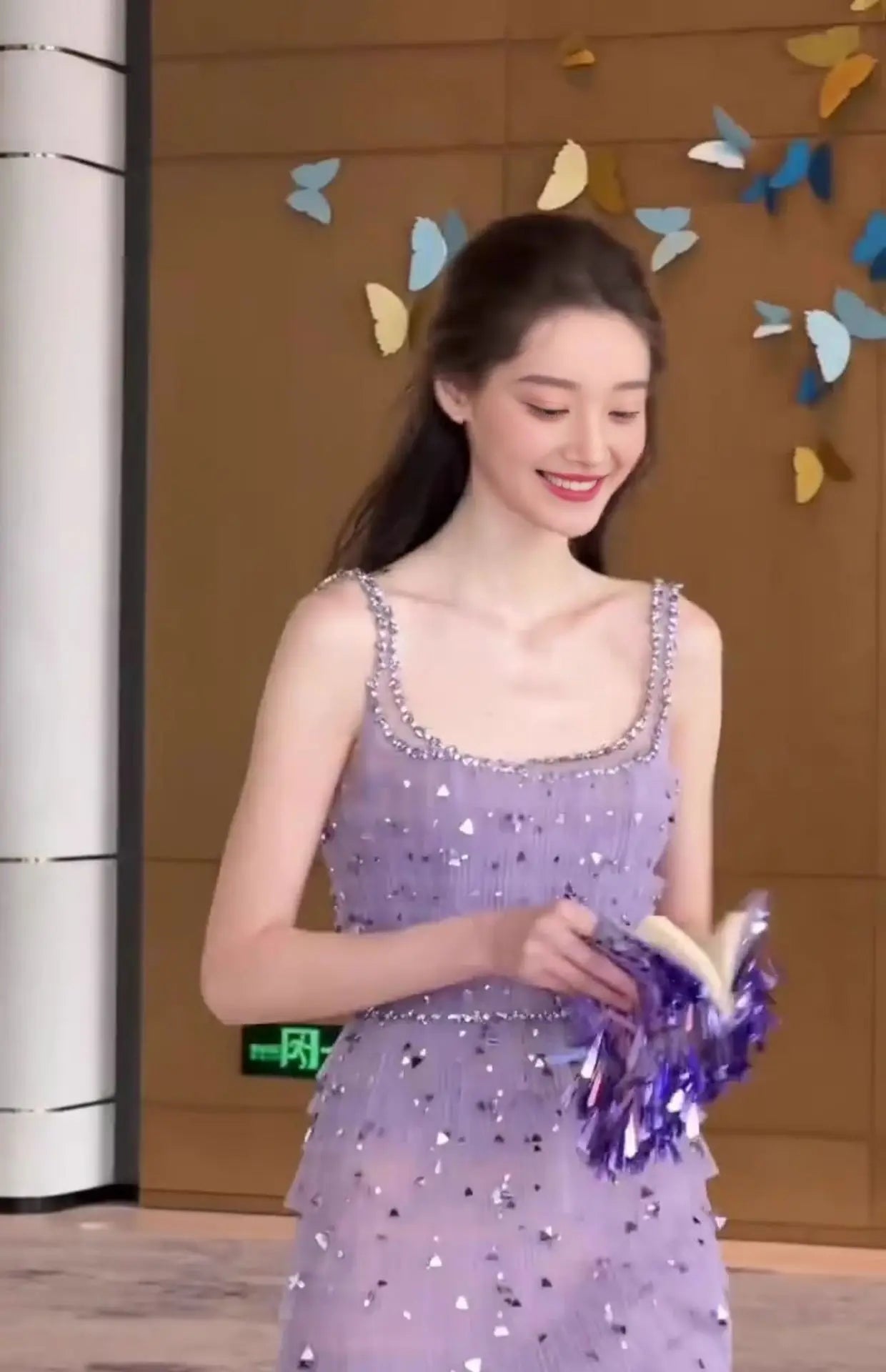 Woman in a purple sequined dress holding a book against a decorative butterfly wall.