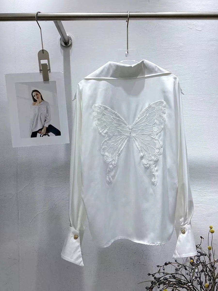 White blouse with butterfly design on a hanger against a white wall.