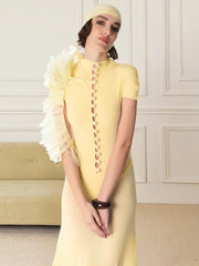Woman wearing a yellow dress with decorative buttons and ruffled sleeves in an elegant room.