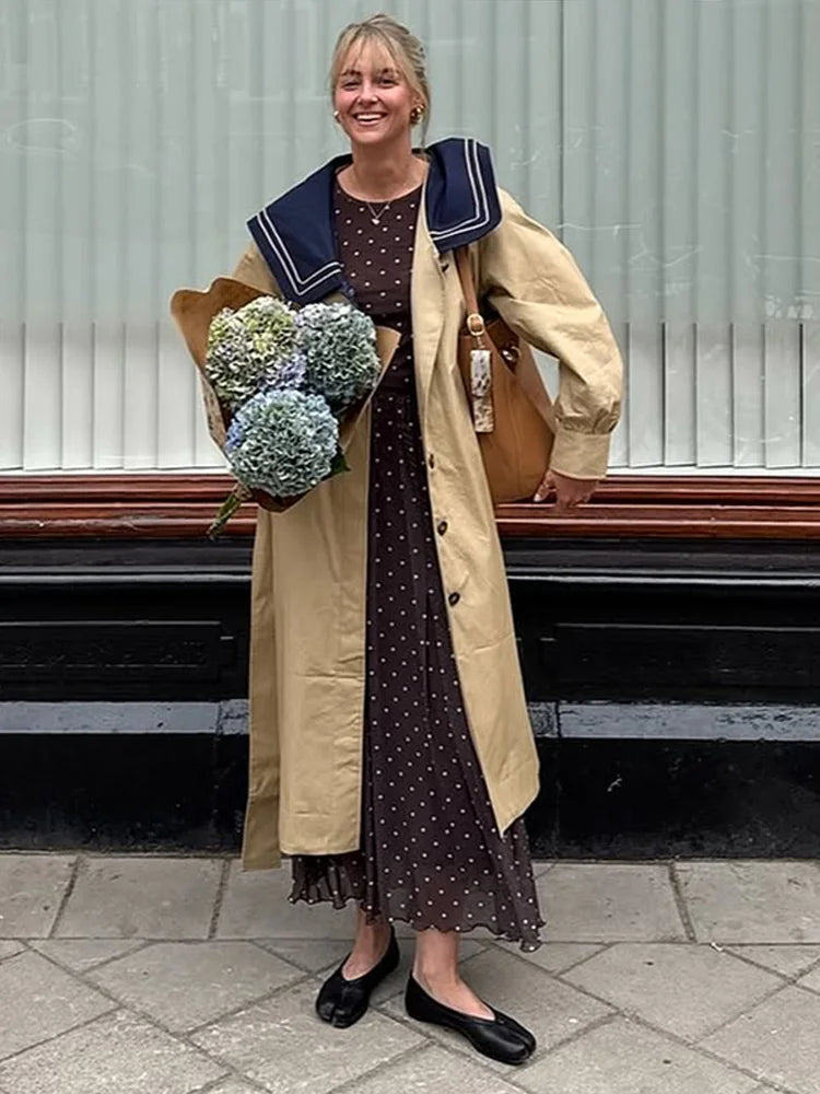 Smiling woman wearing a trench coat with navy sailor collar and polka dot dress, holding flowers