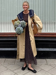 Smiling woman wearing a trench coat with navy sailor collar and polka dot dress, holding flowers