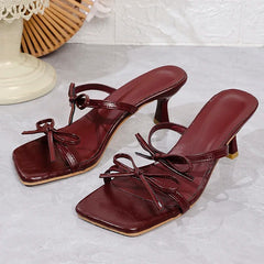 Pair of maroon high-heeled sandals with bow details on a light gray surface.