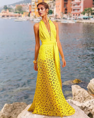 Model wearing a yellow halter-neck maxi dress with floral eyelet cut-outs and a tie waist, standing on rocks by the sea.