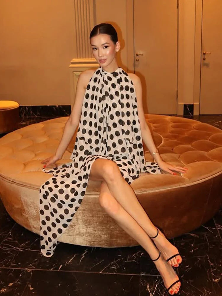 Woman in a polka dot dress sitting on a round brown ottoman in an indoor setting.
