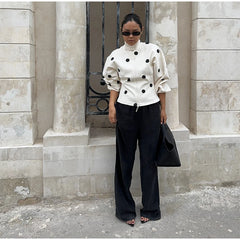 Woman wearing a white top with black polka dots and black pants standing against a stone wall.