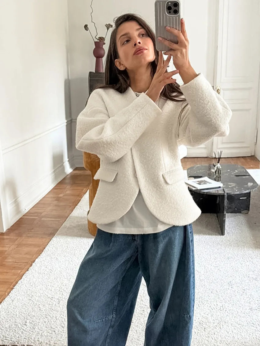 Woman taking a mirror selfie wearing a white blazer and blue jeans in a room with wooden flooring.
